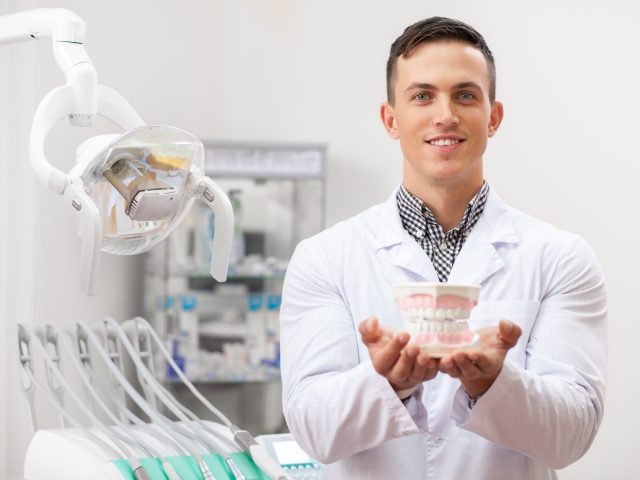 young-handsome-male-dentist-smiling-holding-out-denture-mold-camera